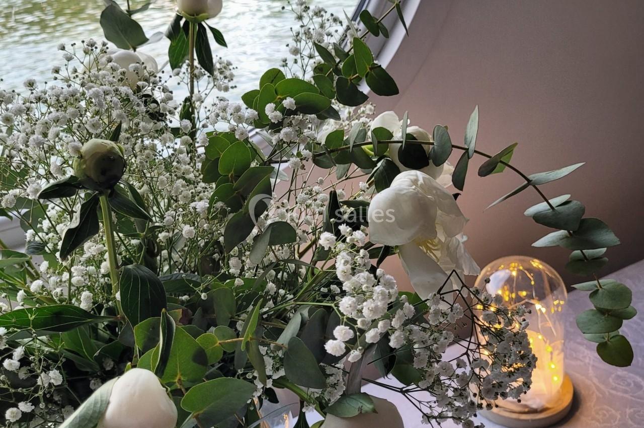 Bouquet de fleurs blanches et feuillages verts posé près d'une fenêtre ronde avec vue sur une rivière.