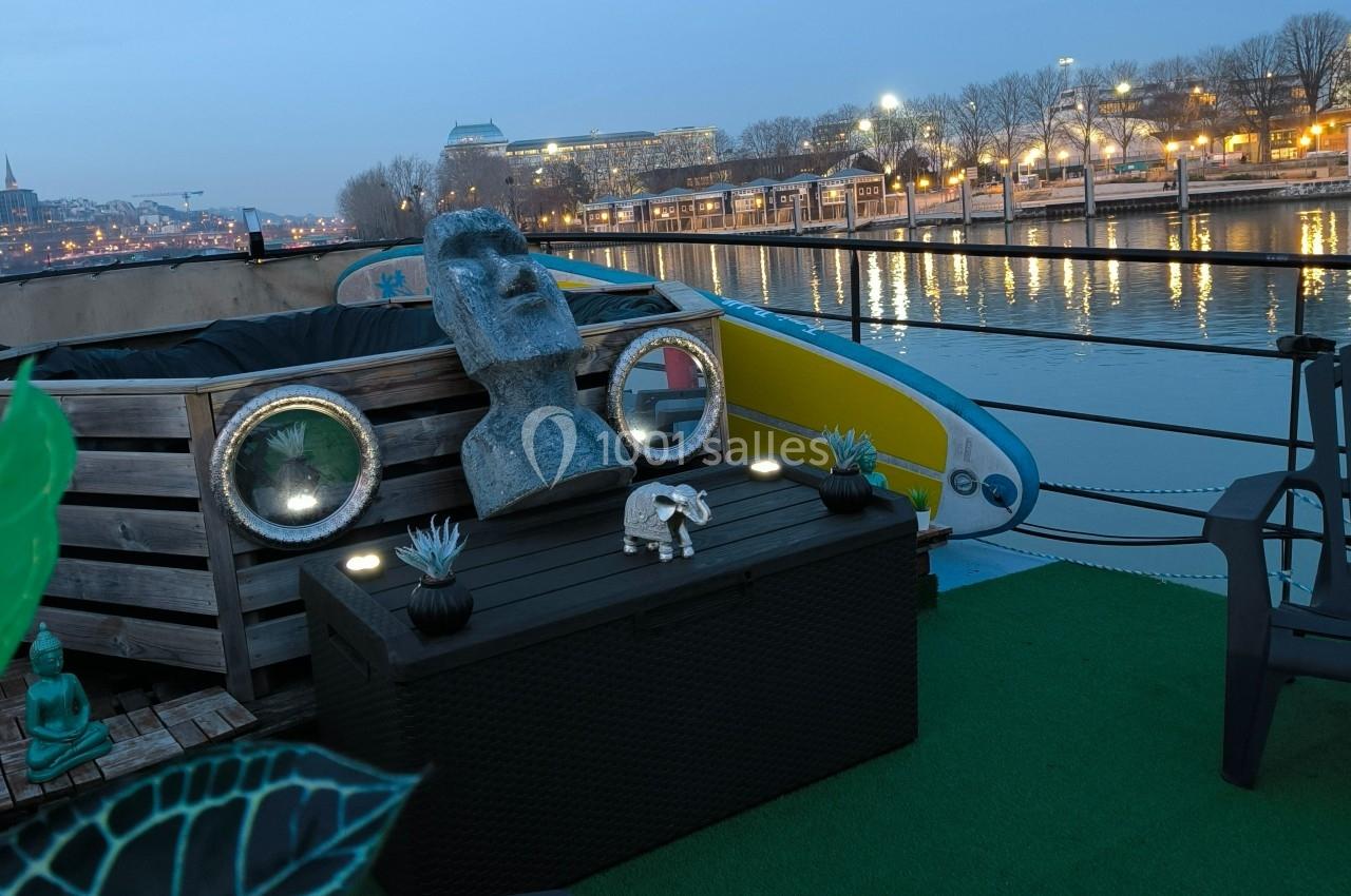 Statue et décorations sur une terrasse de péniche au bord d'un fleuve, avec des lumières et bâtiments en arrière-plan.