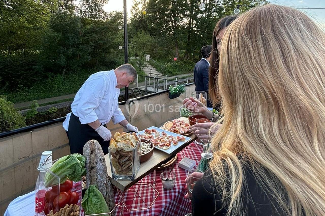 Un chef prépare des amuse-bouches sur une table en plein air, entouré de convives et d'une vue sur un jardin.
