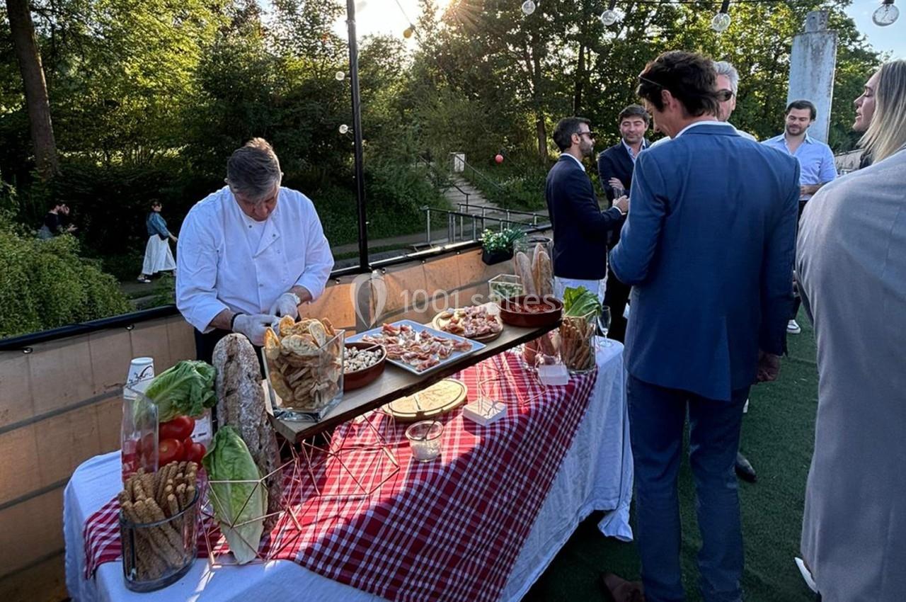 Un chef prépare des plats sur une table garnie de nourriture lors d'un événement en extérieur, entouré de convives.