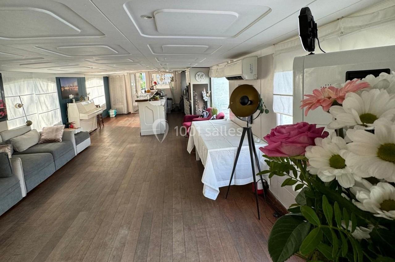 Intérieur lumineux d'une péniche aménagée avec des canapés, une table dressée et des fleurs au premier plan.