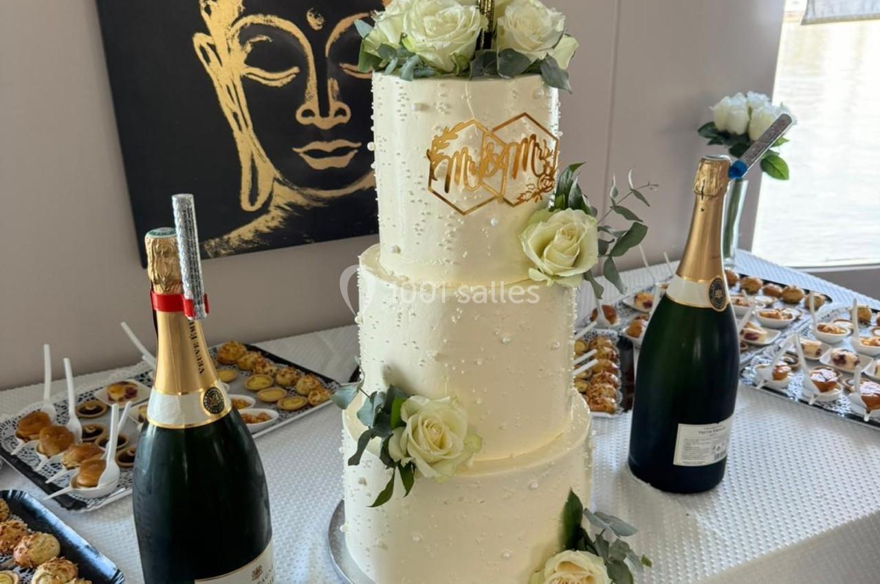 Gâteau de mariage à trois étages décoré de roses blanches, entouré de champagne et de desserts sur une table.
