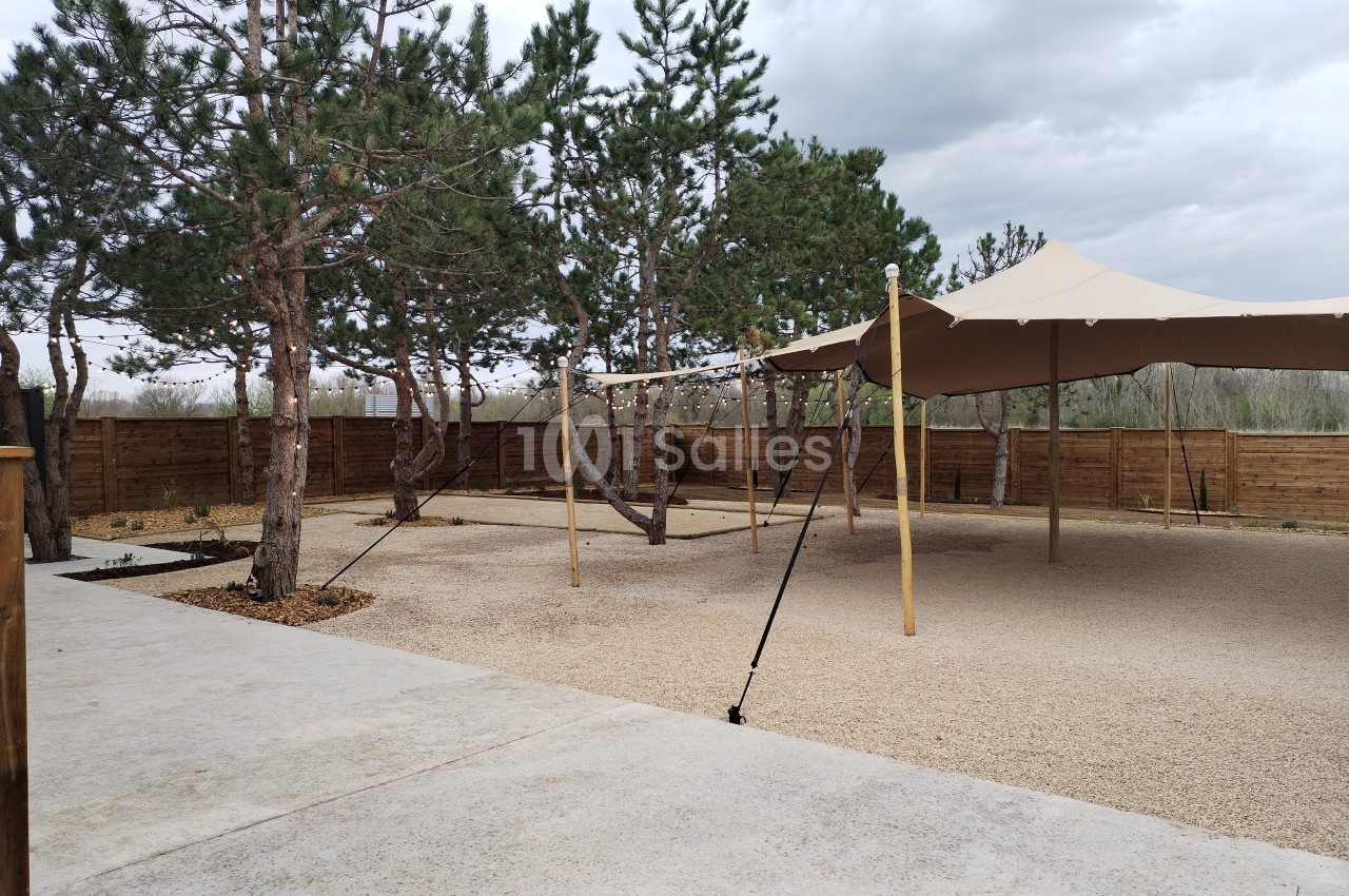 Espace extérieur avec sol en gravier, arbres, tente beige tendue et clôture en bois sous un ciel nuageux.