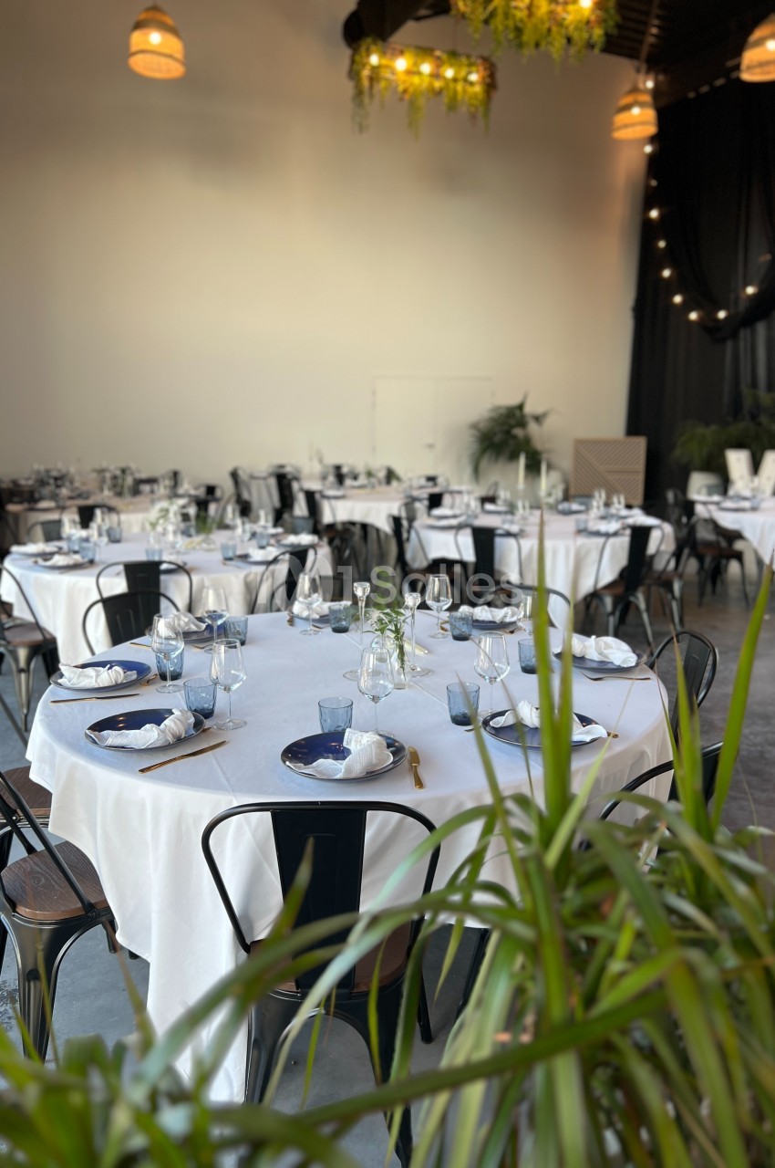 Salle de réception décorée avec des tables rondes dressées, nappes blanches, chaises noires et éclairage chaleureux.
