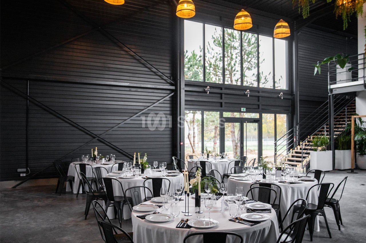 Salle de réception moderne avec tables rondes dressées, grandes baies vitrées et décoration végétale suspendue.