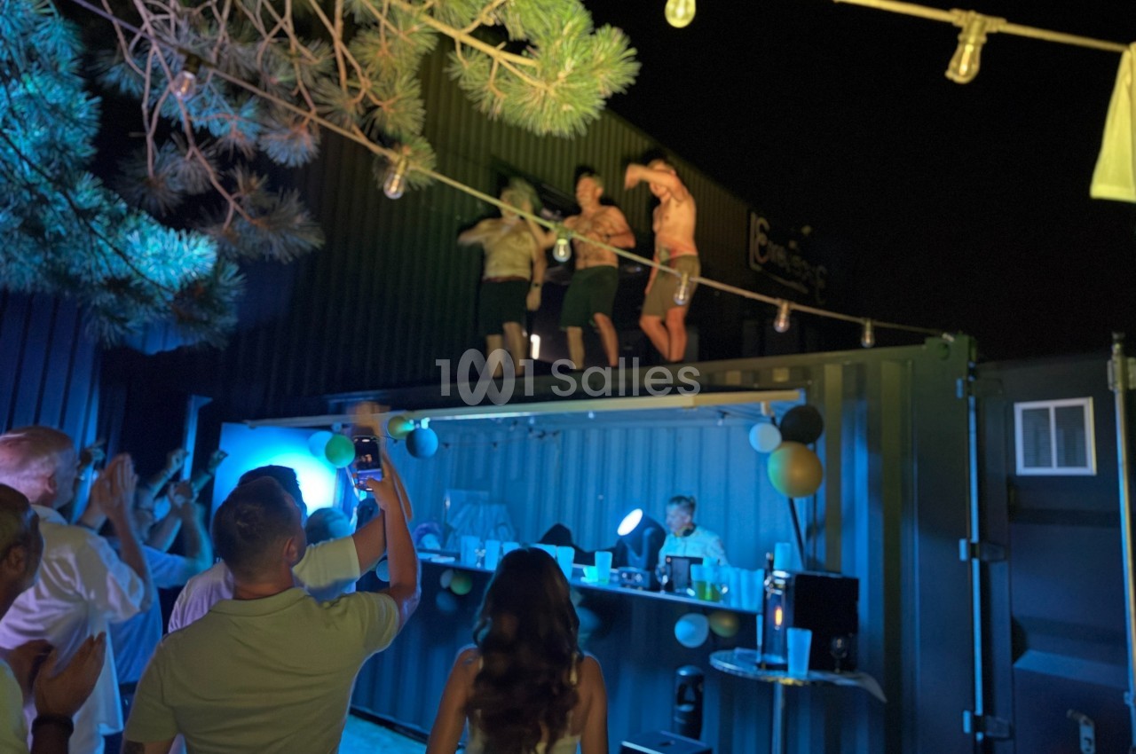 Des personnes dansent sur le toit d'un conteneur éclairé, entouré de spectateurs lors d'une fête nocturne.