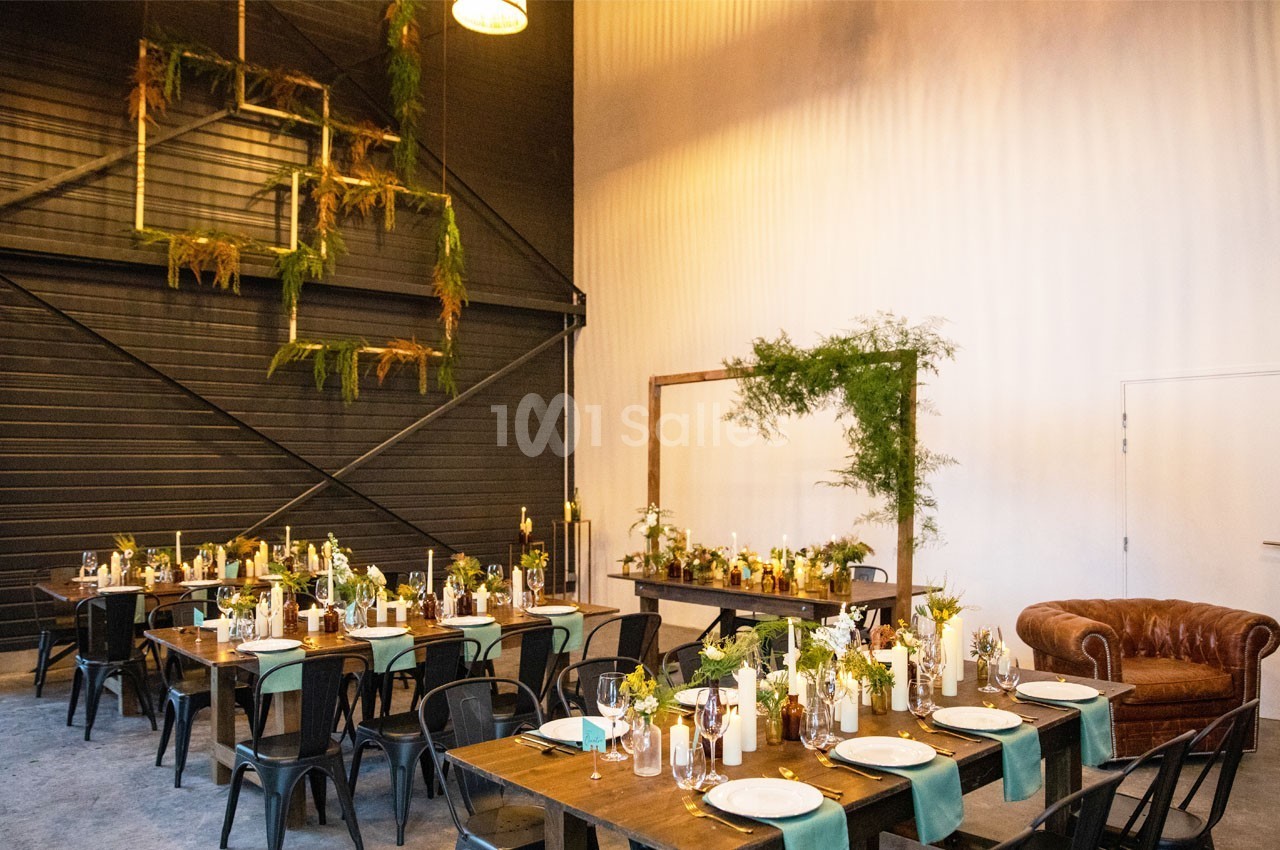 Salle décorée pour un événement, avec tables en bois, chaises noires, plantes suspendues et éclairage chaleureux.