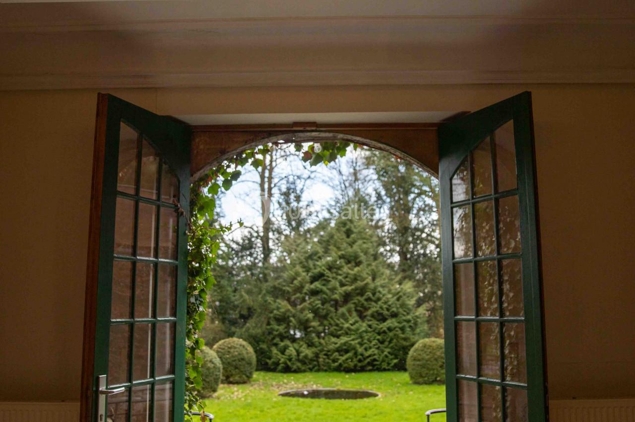 Porte ouverte donnant sur un jardin verdoyant avec pelouse, arbustes taillés et bassin central, éclairage au plafond.