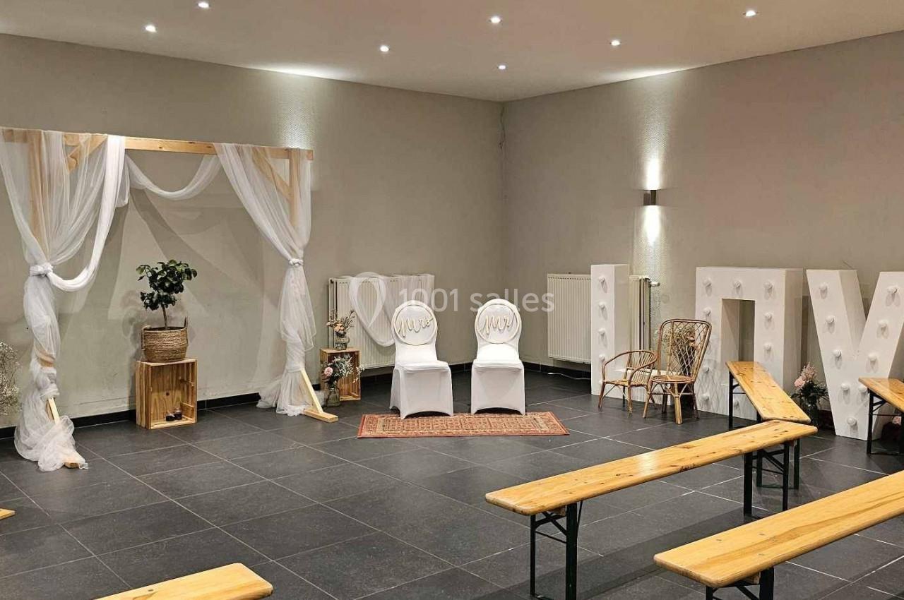 Salle décorée pour une cérémonie avec arche en bois et tissu, chaises blanches, bancs en bois et lettres lumineuses ’LOVE’.