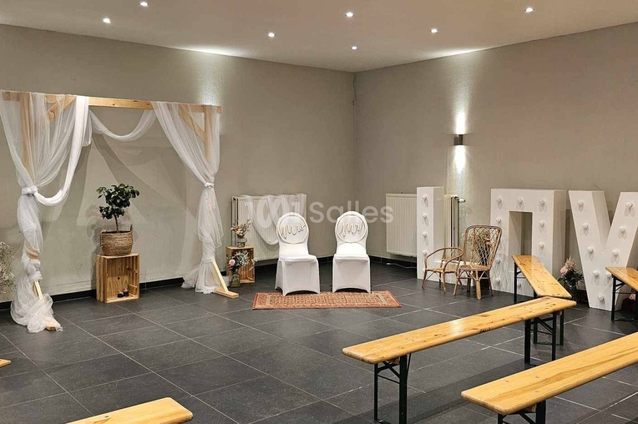 Salle décorée pour une cérémonie avec arche en bois et tissu, chaises blanches, bancs en bois et lettres lumineuses ’LOVE’.