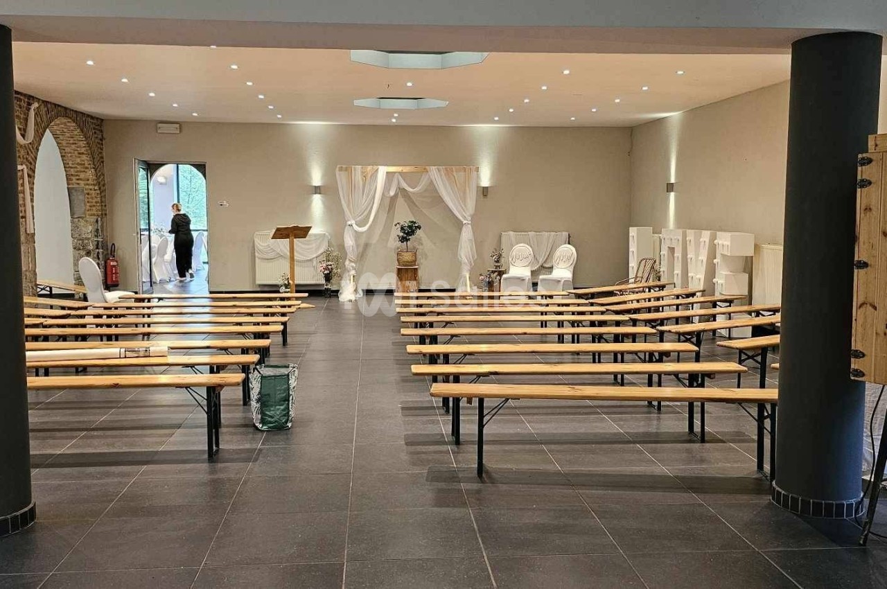 Salle de réception avec bancs en bois alignés, décorations blanches et colonnes noires, vue vers une entrée lumineuse.