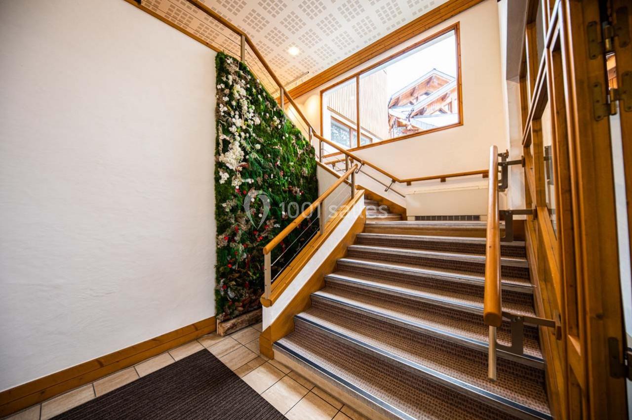 Escalier intérieur en bois avec une rampe, un mur végétal et une grande fenêtre laissant entrer la lumière naturelle.