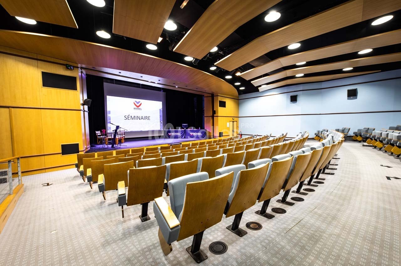Salle de conférence avec rangées de sièges vides, scène équipée d'un écran affichant ’Séminaire’.