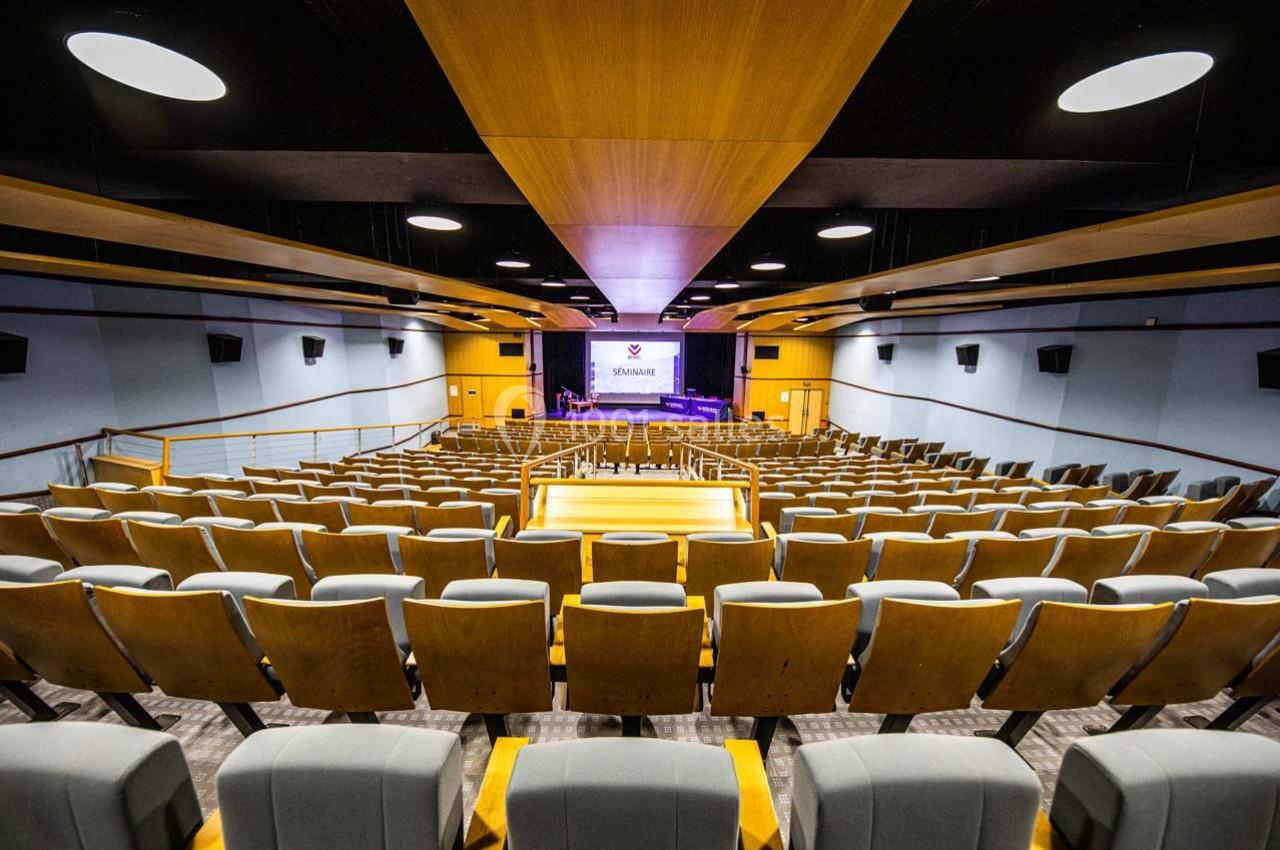 Salle de conférence vide avec rangées de sièges en bois, scène équipée d'un écran de projection et éclairage au plafond.