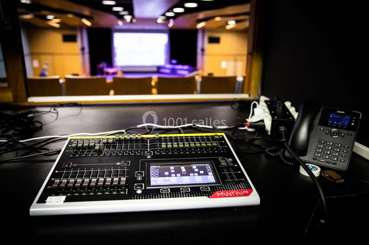 Table de mixage lumineuse dans une régie technique, avec vue sur une salle de conférence et un écran de projection.