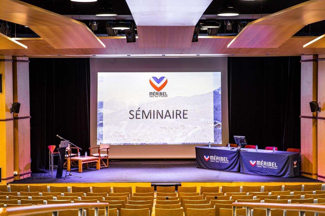 Salle de conférence avec scène, écran affichant ’Séminaire’ et tables équipées pour une présentation.