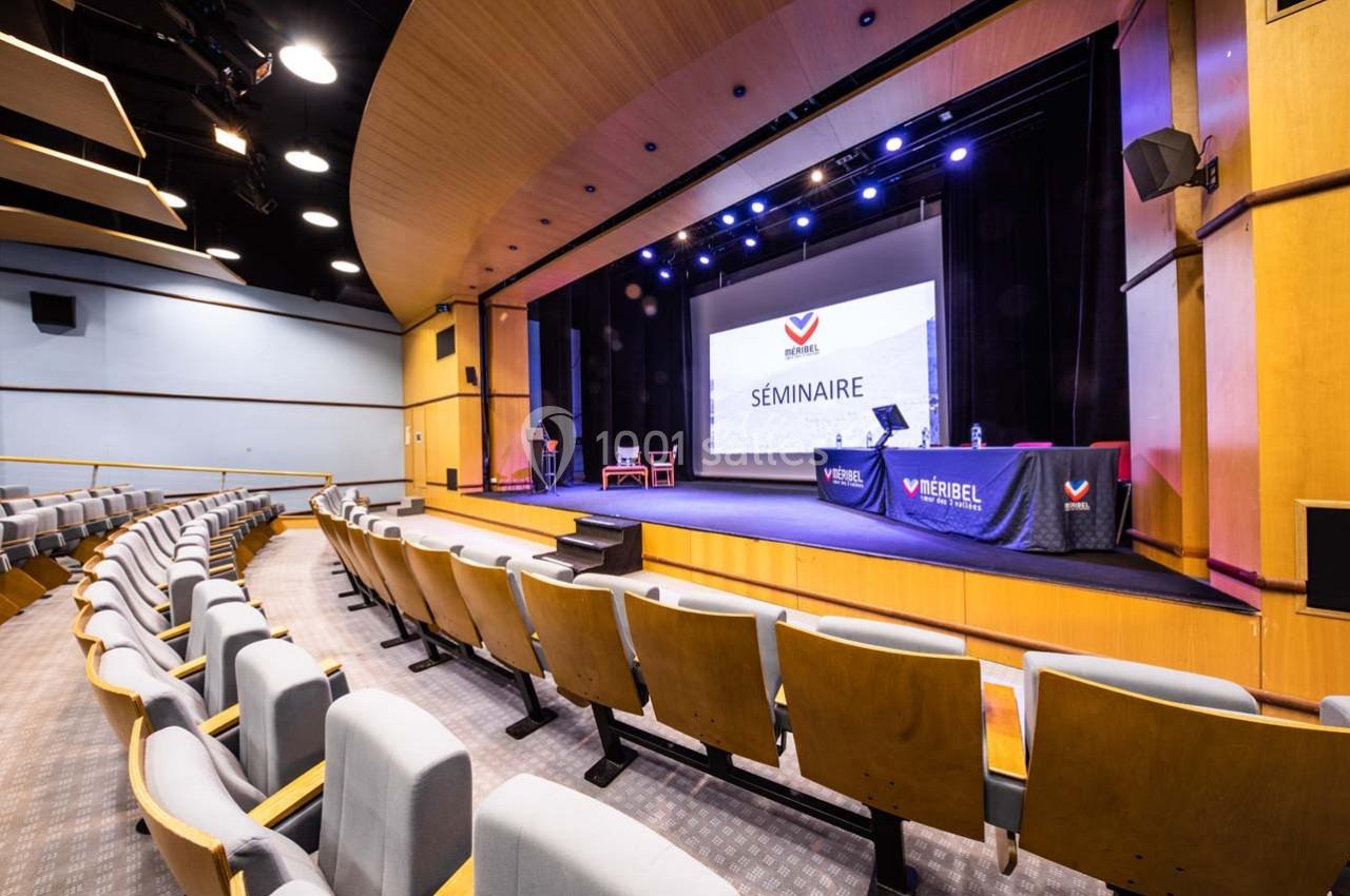 Salle de conférence vide avec un écran de projection affichant ’Séminaire’, des tables et des chaises sur la scène.
