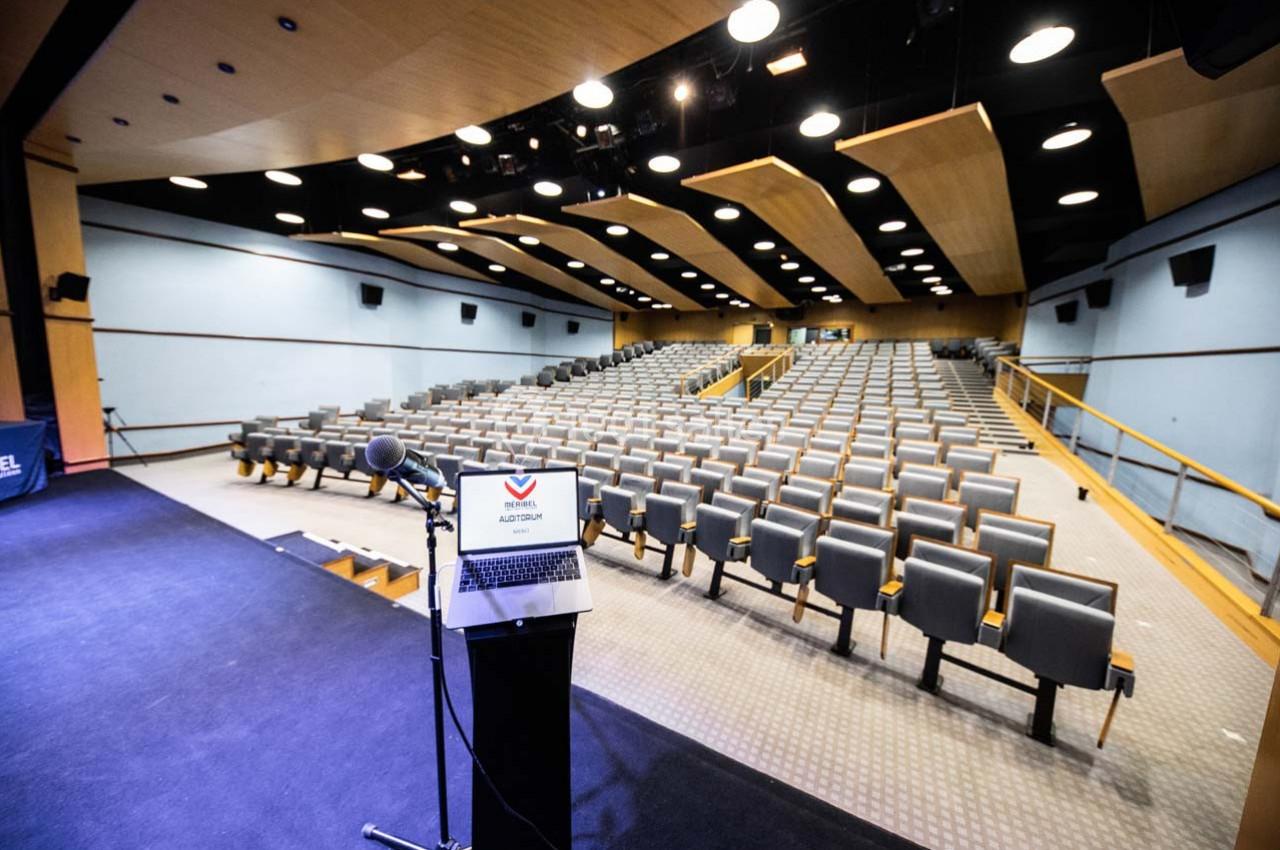 Salle de conférence vide avec rangées de sièges gris, vue depuis un pupitre équipé d'un micro et d'un ordinateur portable.
