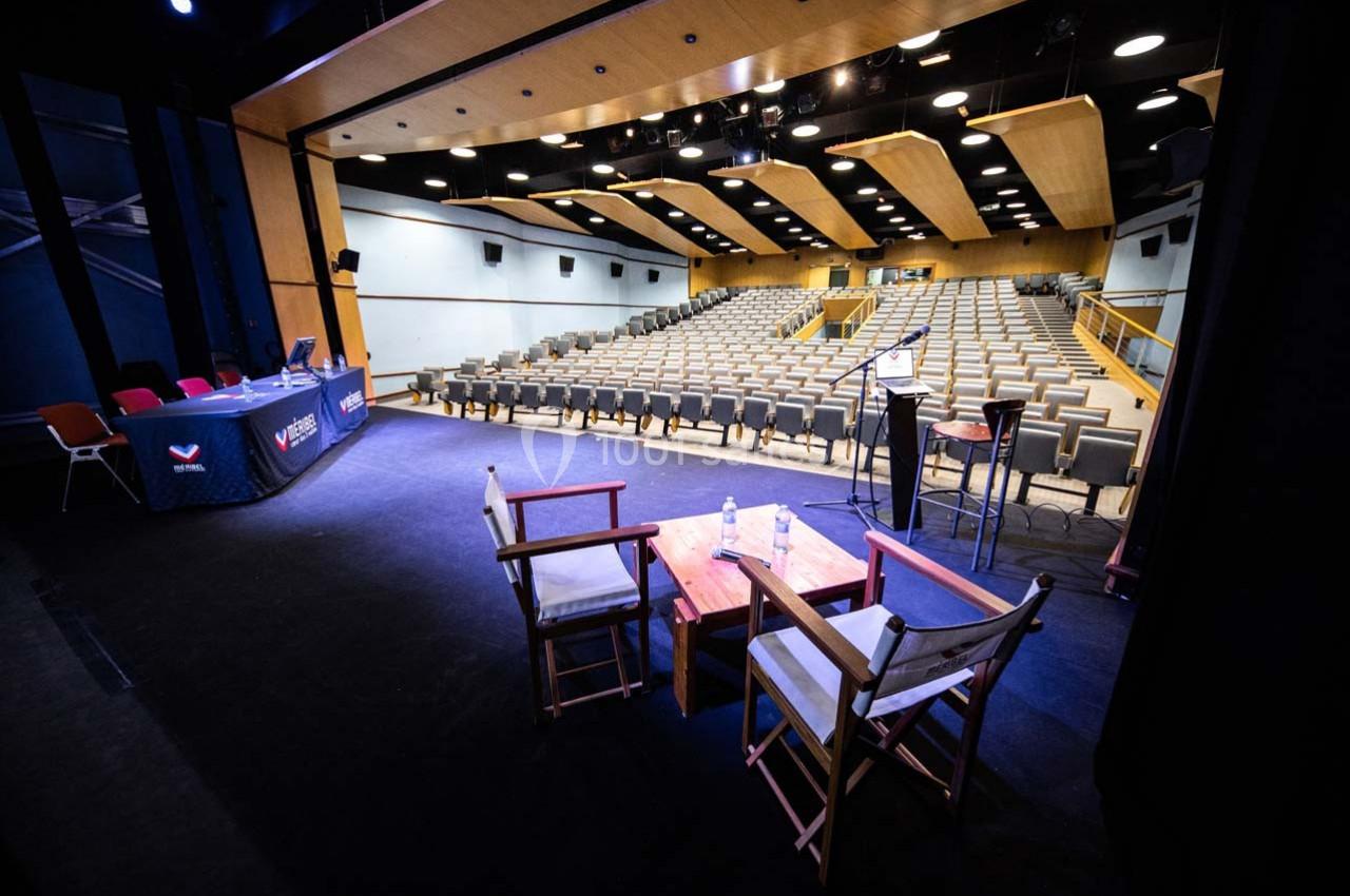 Salle de conférence vide avec rangées de sièges, scène équipée de chaises, table et micros sous un éclairage artificiel.