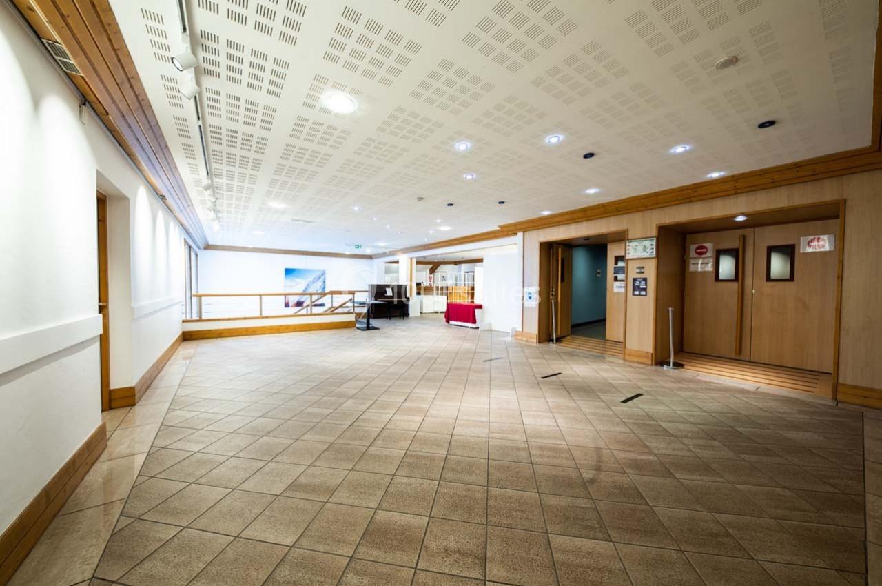 Couloir lumineux avec sol carrelé, murs blancs, plafond acoustique et deux ascenseurs en bois à droite.
