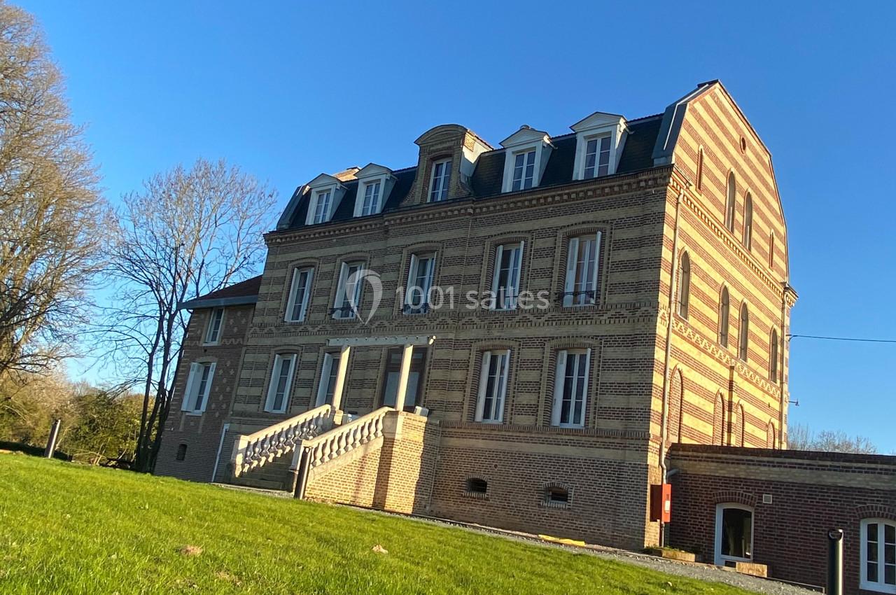 Bâtiment en briques à trois étages avec toit mansardé, entouré de pelouse et d'arbres sous un ciel bleu.