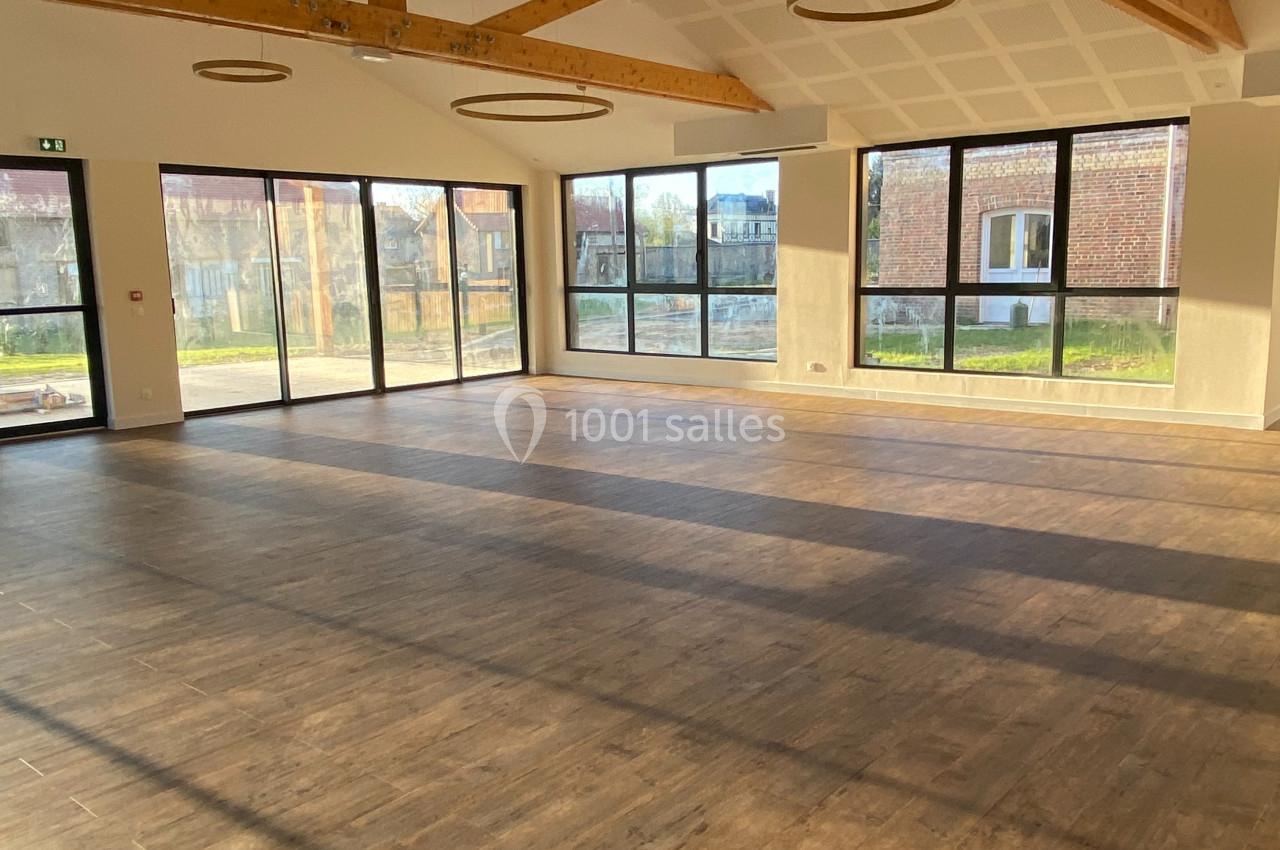 Salle vide avec parquet en bois, grandes baies vitrées laissant entrer la lumière naturelle et poutres apparentes au plafond.