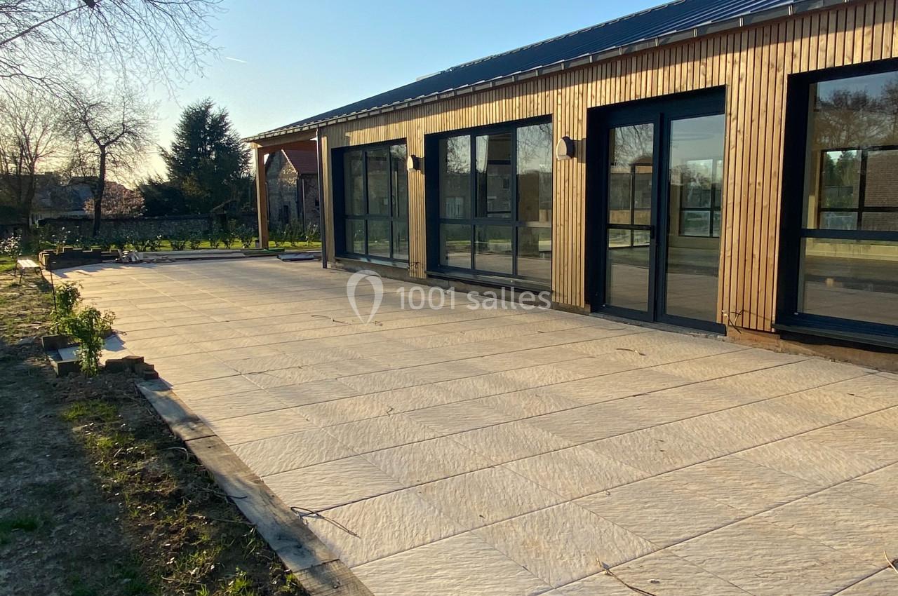 Terrasse en dalles claires devant une maison moderne en bois avec grandes baies vitrées, sous un ciel dégagé.