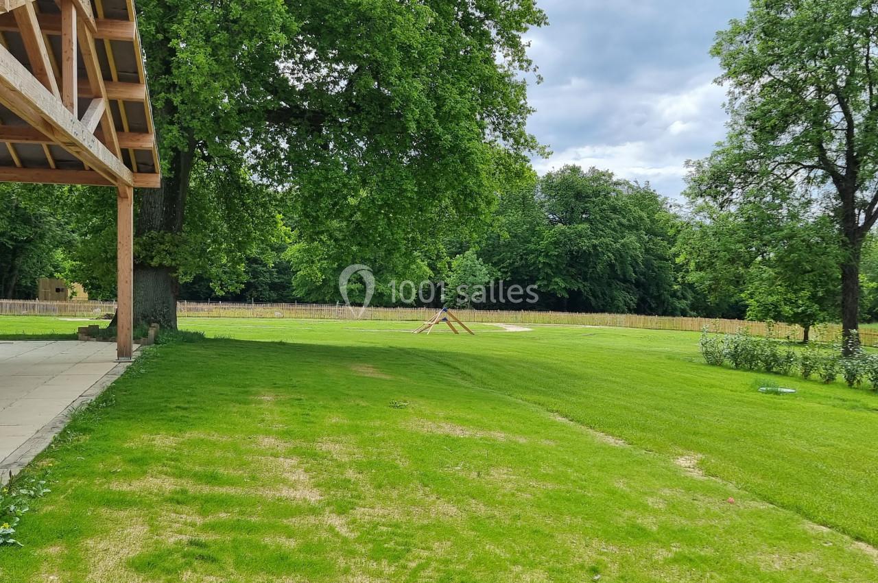 Vue d'un jardin verdoyant avec pelouse, arbres et une structure en bois sur la gauche sous un ciel partiellement nuageux.