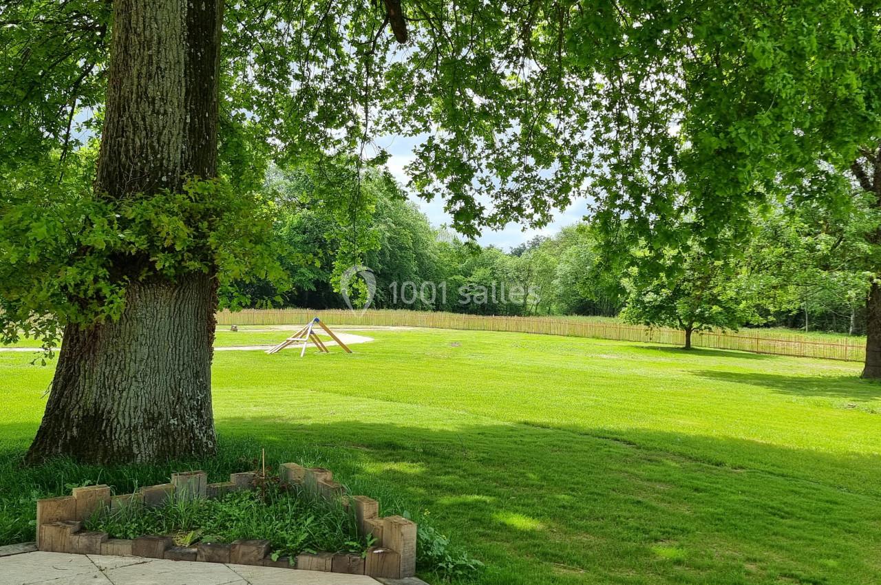 Un grand arbre ombrage un jardin verdoyant avec une structure en bois au loin, entouré de nature.