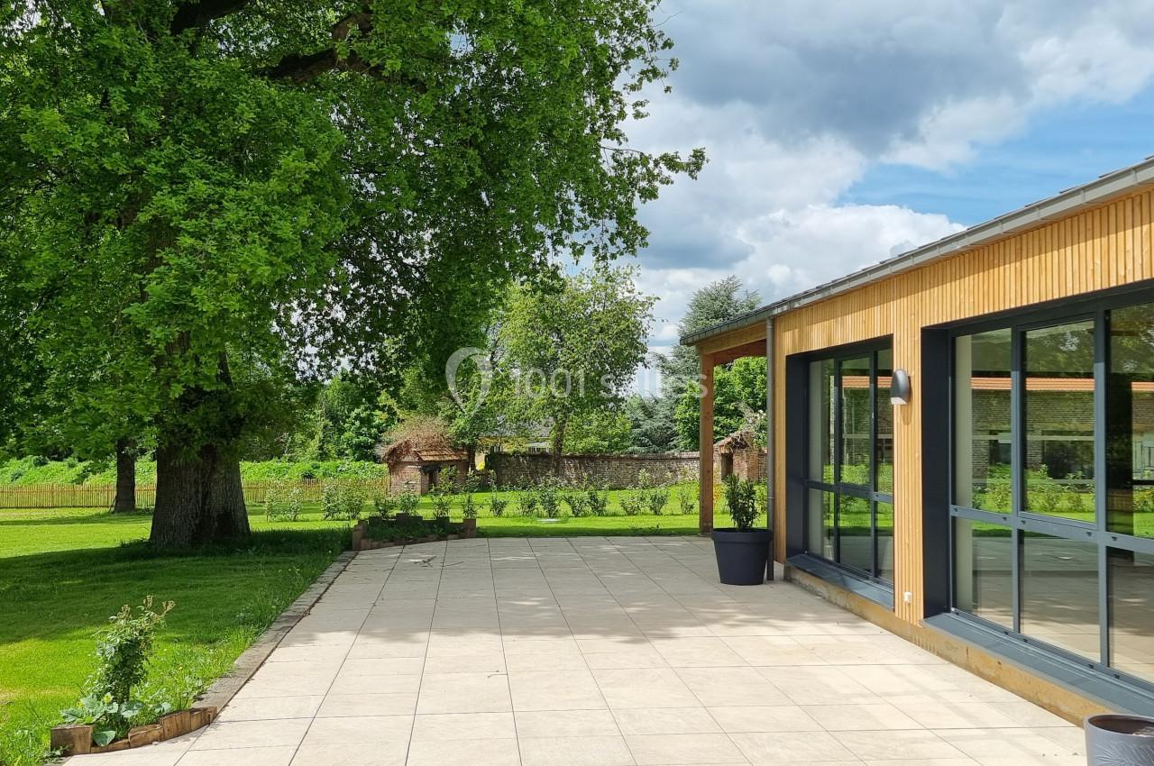 Terrasse en dalles claires bordant un bâtiment en bois avec de grandes baies vitrées, entourée d'un jardin verdoyant.