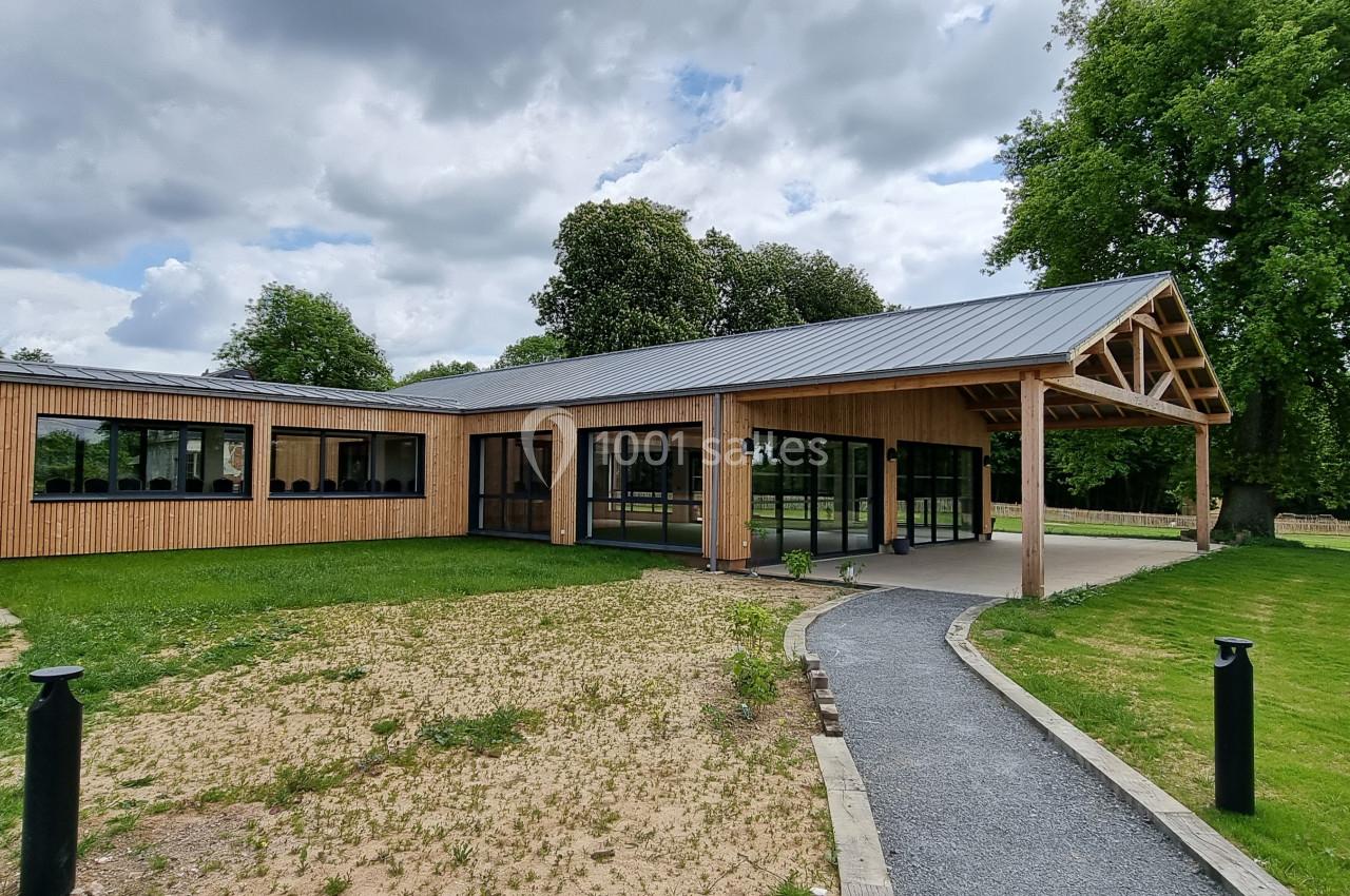 Bâtiment moderne en bois avec toit en métal, grandes baies vitrées, entouré de pelouse et d'arbres sous un ciel nuageux.
