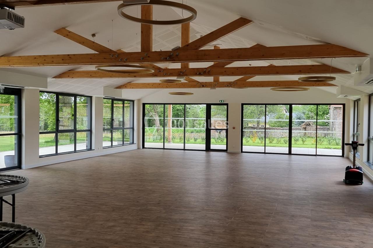Salle lumineuse avec parquet, grandes baies vitrées donnant sur un espace vert, et poutres apparentes au plafond.
