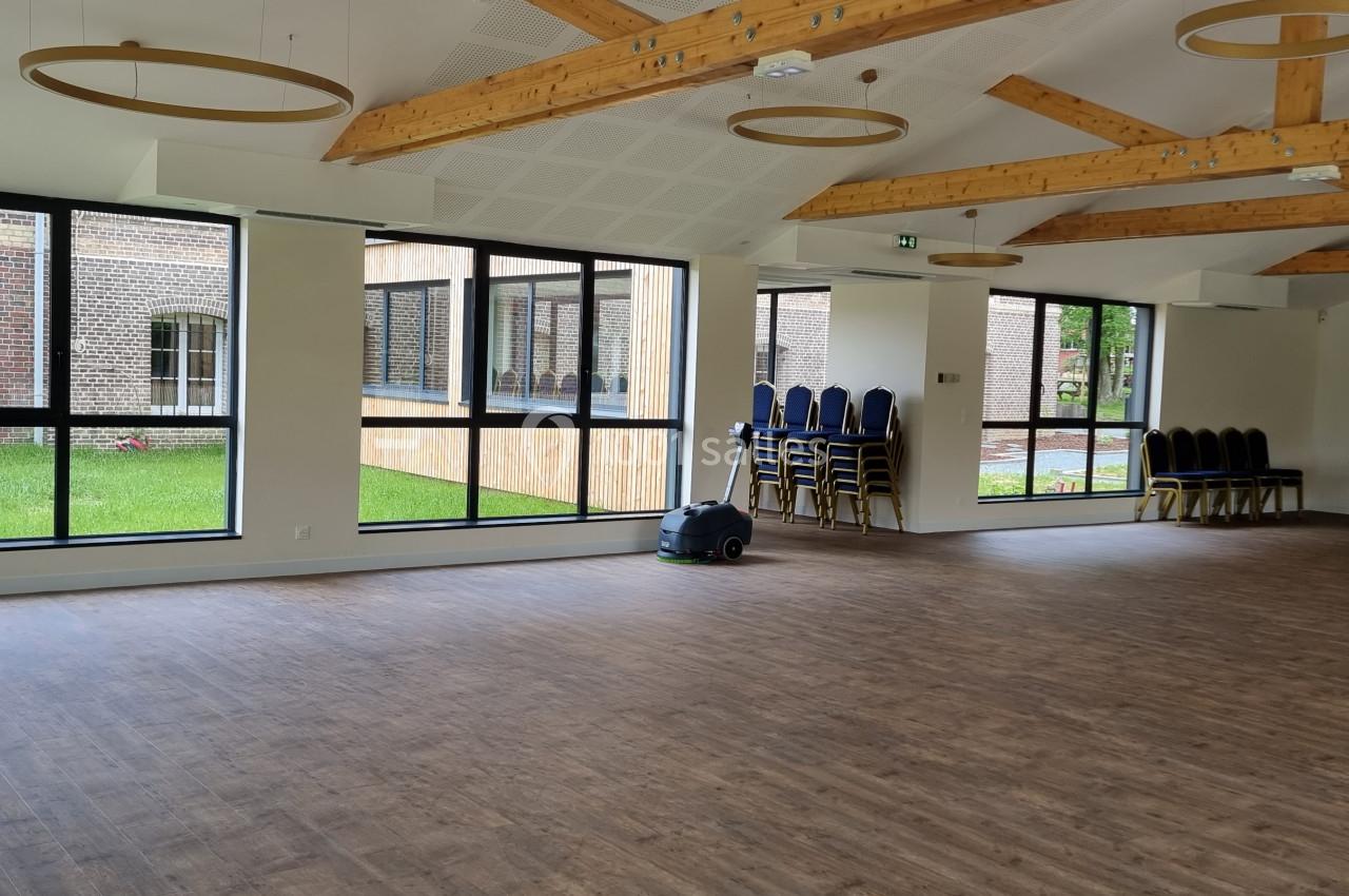 Salle lumineuse avec parquet, poutres apparentes, grandes fenêtres donnant sur un espace vert, chaises empilées sur le côté.