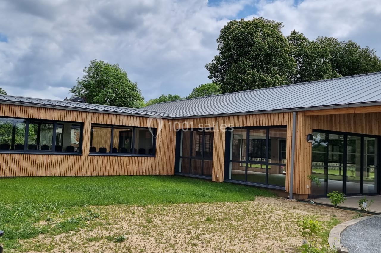 Bâtiment moderne en bois avec de grandes baies vitrées, entouré de pelouse et d'arbres sous un ciel partiellement nuageux.