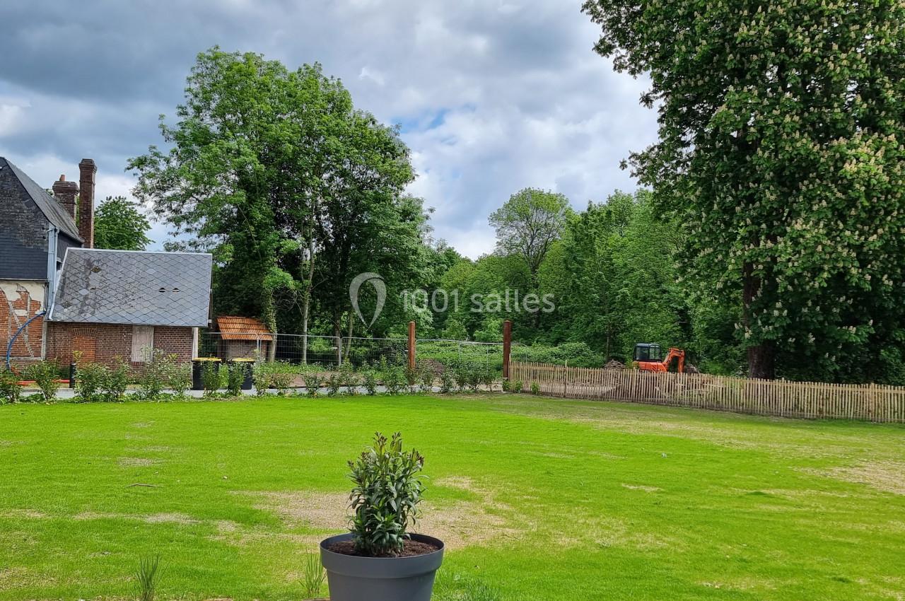 Jardin avec pelouse, arbres, clôture en bois et une maison en arrière-plan sous un ciel partiellement nuageux.