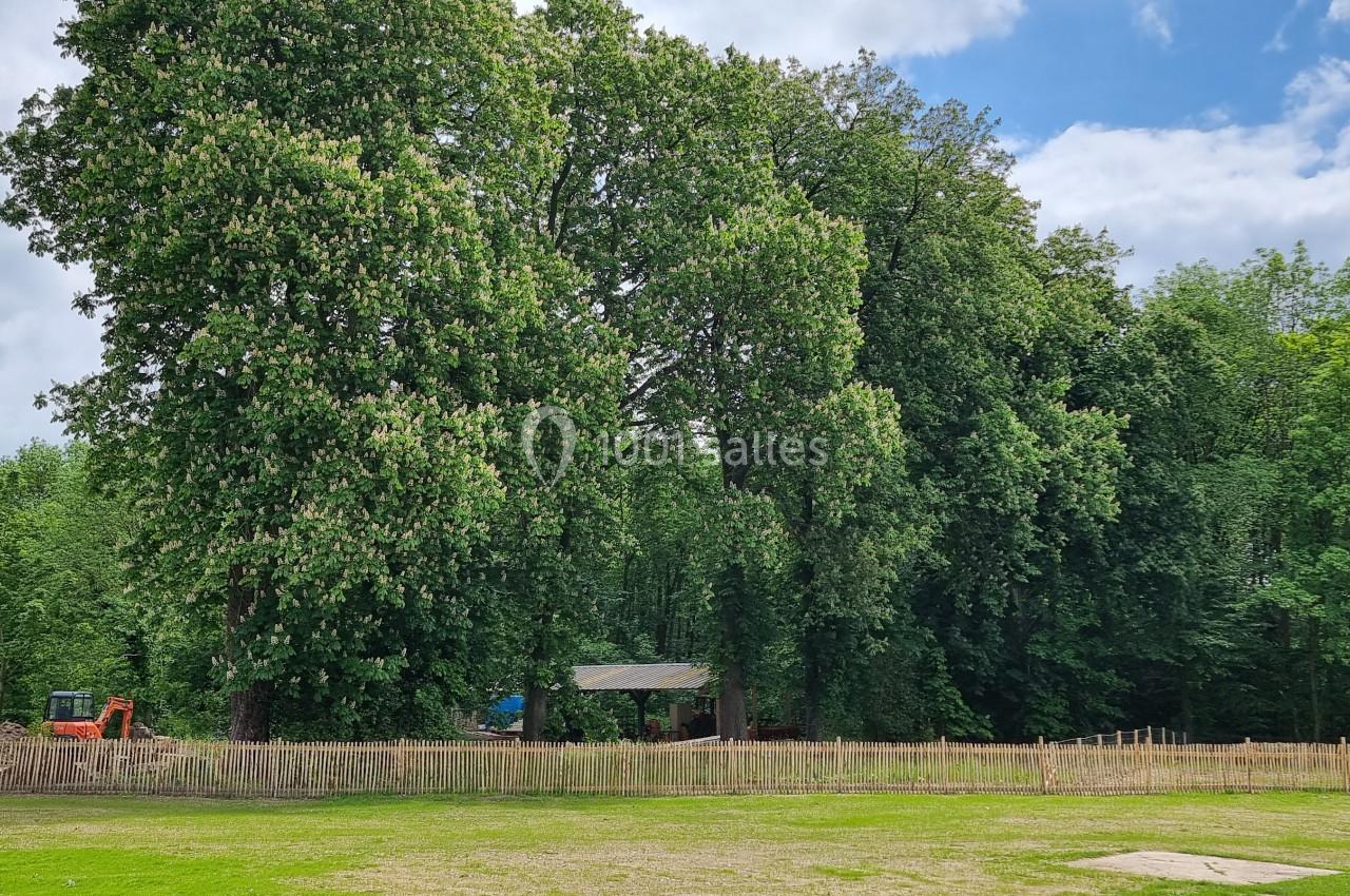 Grandes arbres bordant une clairière avec une clôture en bois, une petite structure et une pelouse au premier plan.