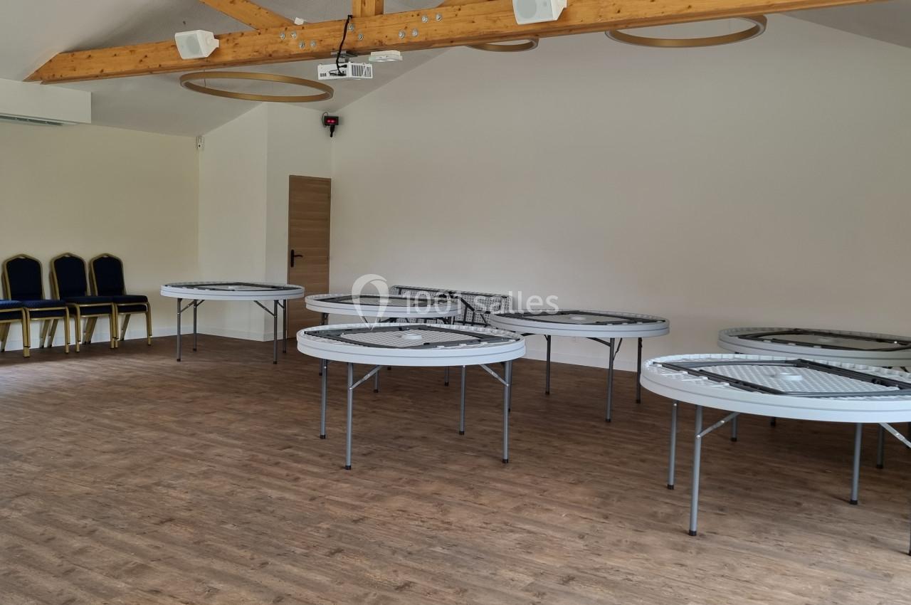 Salle lumineuse avec parquet, tables rondes pliantes disposées, chaises empilées près d'une fenêtre et poutres apparentes au…