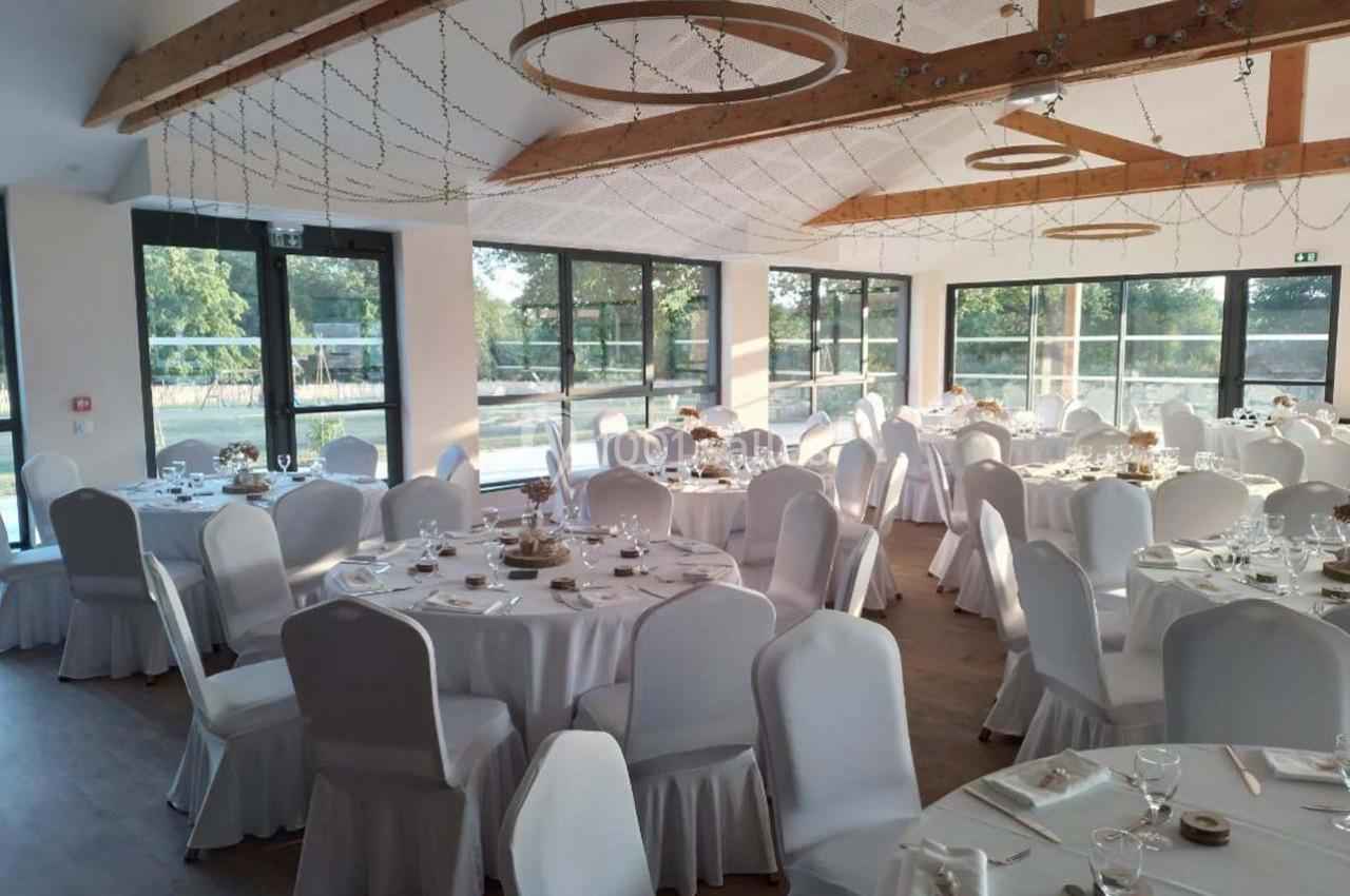 Salle de réception lumineuse avec tables rondes dressées, nappes blanches et grandes baies vitrées donnant sur l'extérieur.