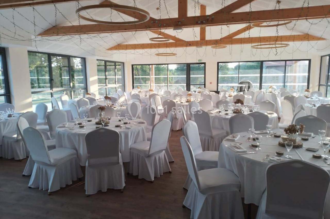Salle de réception lumineuse avec tables rondes dressées, nappes blanches et décorations suspendues en bois et guirlandes.
