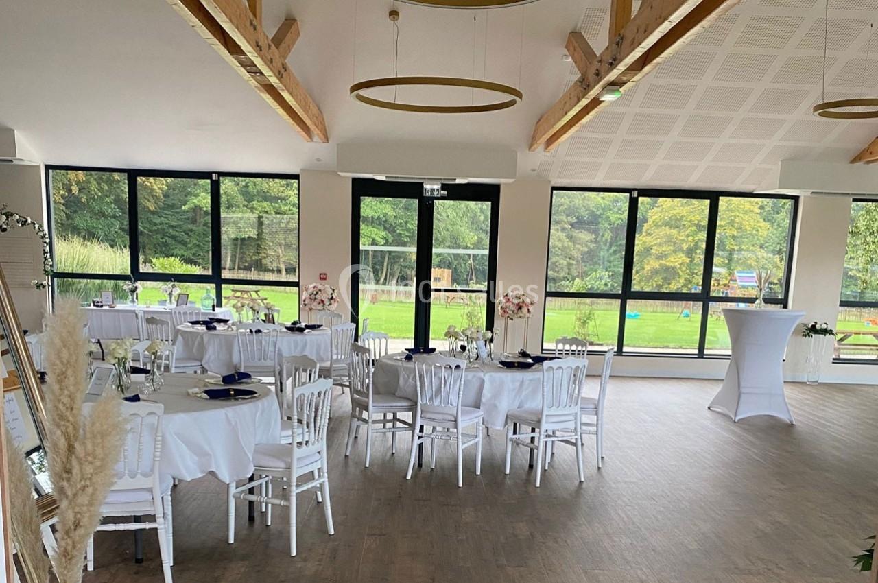 Salle de réception lumineuse avec tables rondes dressées, grandes baies vitrées et vue sur un jardin verdoyant.