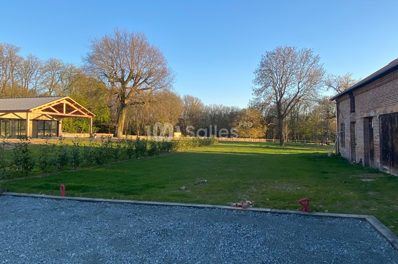 Vue d'un jardin avec pelouse, graviers, bâtiment en briques et structure en bois, entouré d'arbres sous un ciel dégagé.
