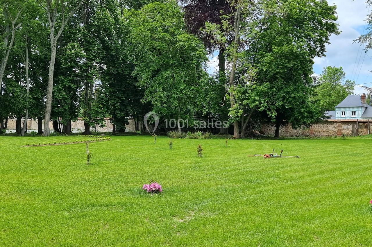 Pelouse verdoyante avec quelques fleurs colorées, entourée d'arbres et d'un mur en pierre, maison visible en arrière-plan.