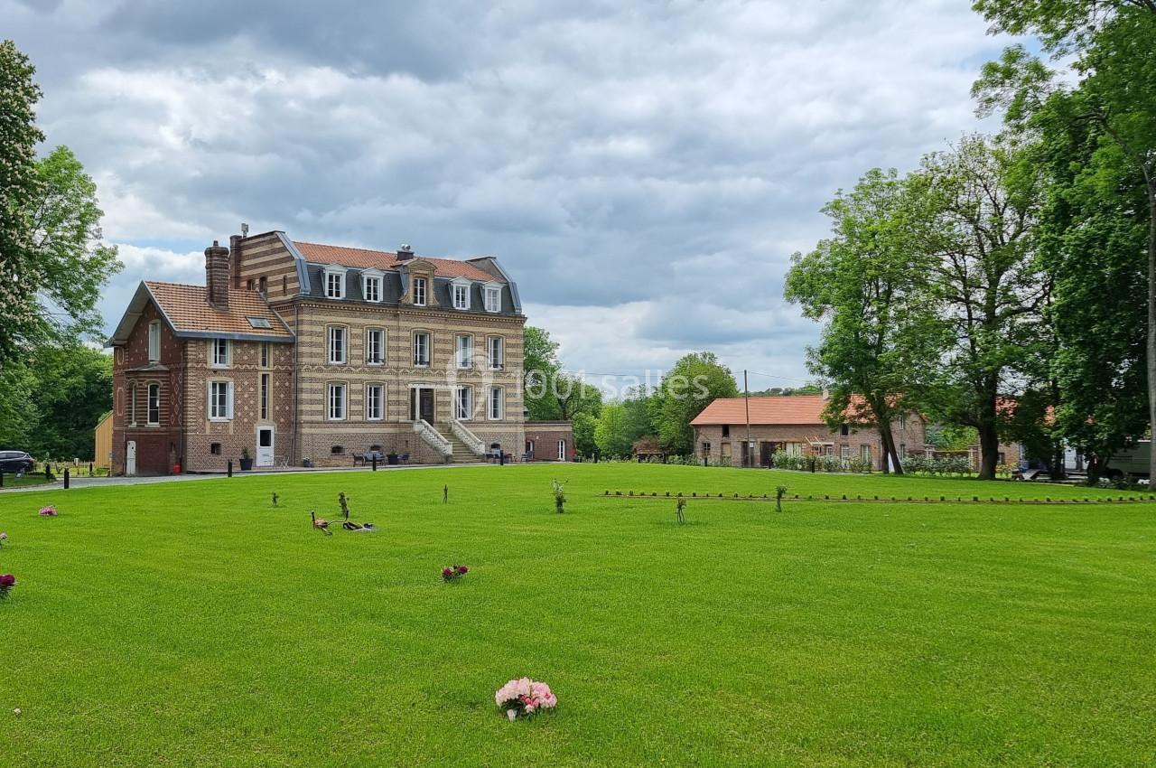 Grand bâtiment en briques entouré d'un vaste jardin verdoyant avec quelques fleurs et arbres sous un ciel nuageux.