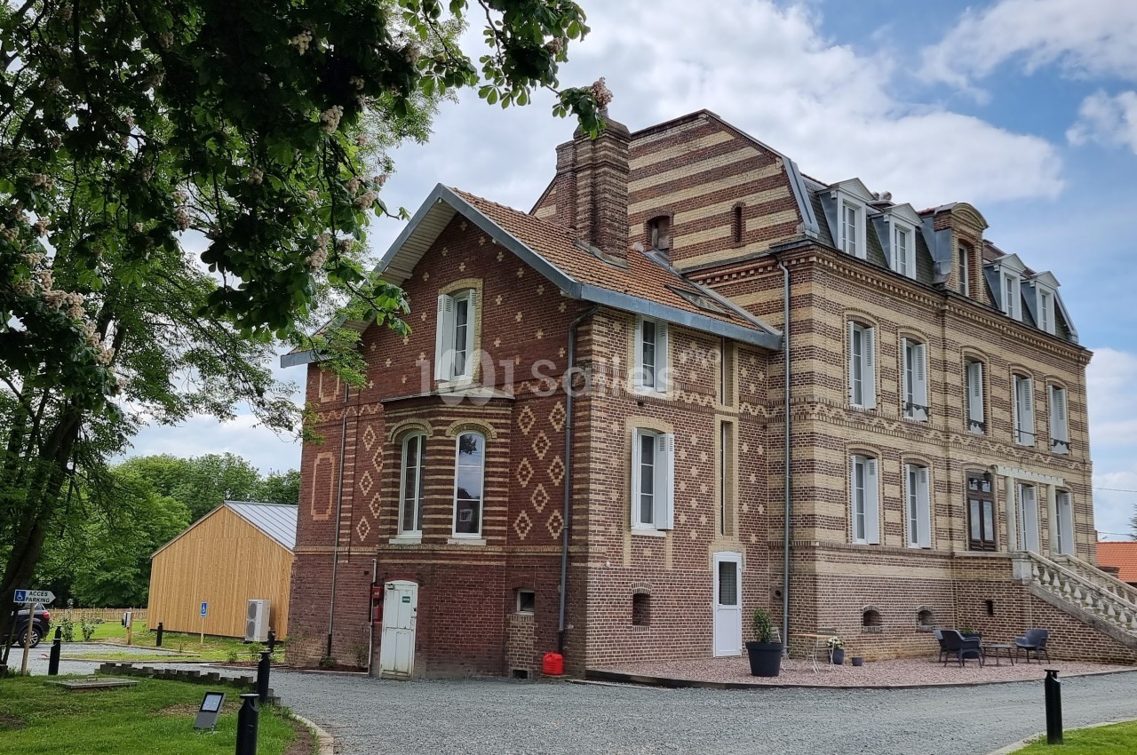 Bâtiment en briques rouges et beiges avec toit mansardé, entouré de verdure et d'un ciel partiellement nuageux.