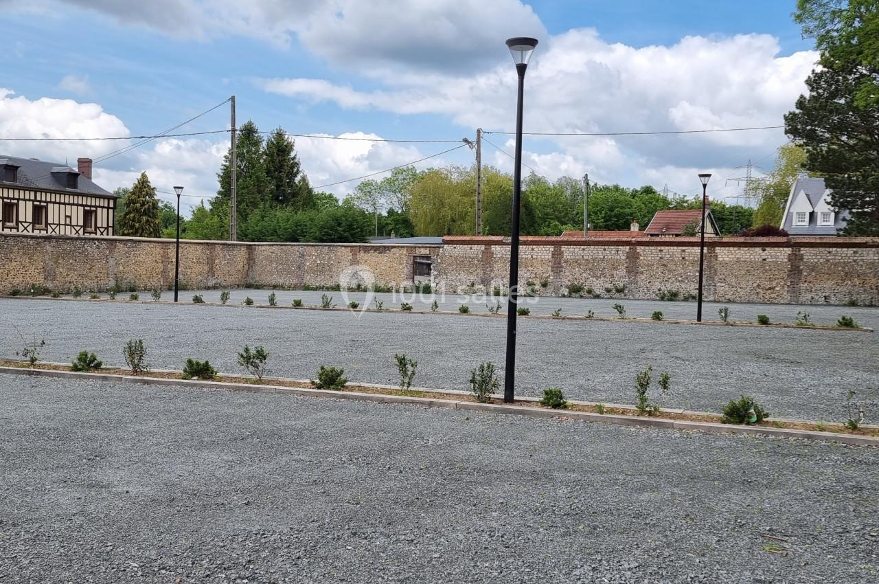 Parking en gravier avec quelques jeunes arbustes, entouré de murs en pierre et bordé de maisons et d'arbres.