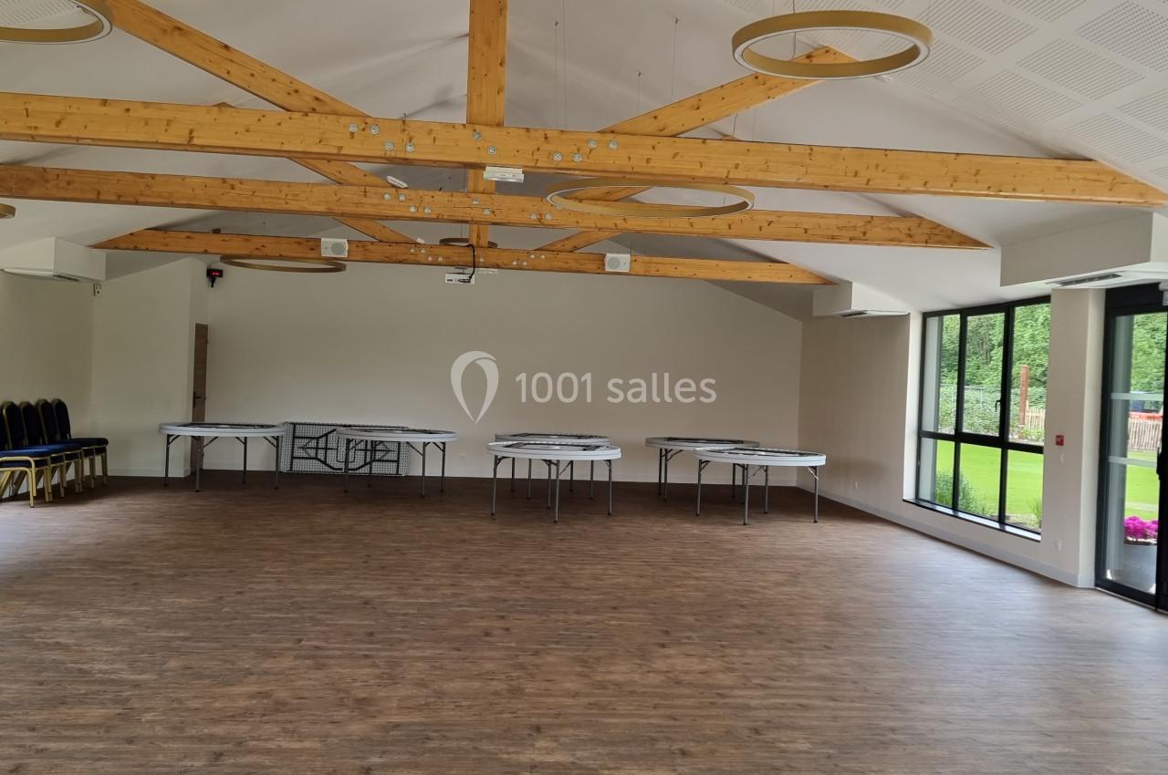 Salle lumineuse avec parquet, poutres apparentes, tables pliées contre le mur et grandes fenêtres donnant sur un jardin.
