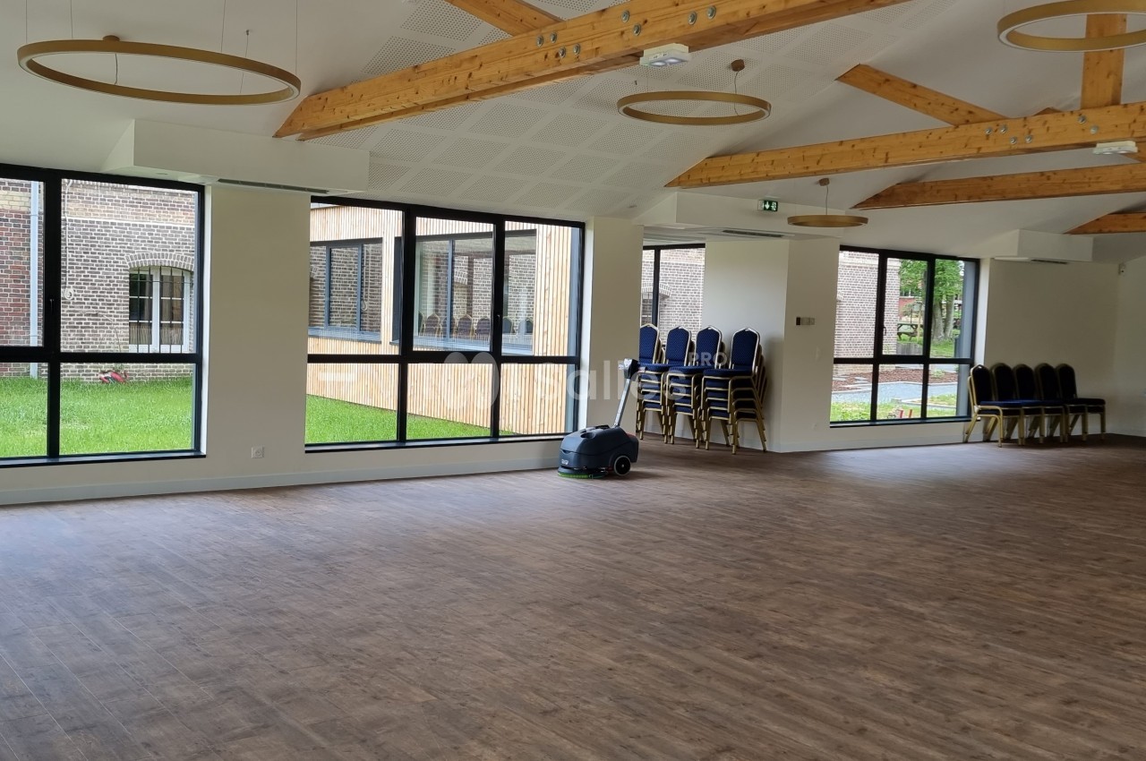 Salle lumineuse avec parquet, grandes fenêtres donnant sur un jardin, poutres apparentes et chaises empilées dans un coin.