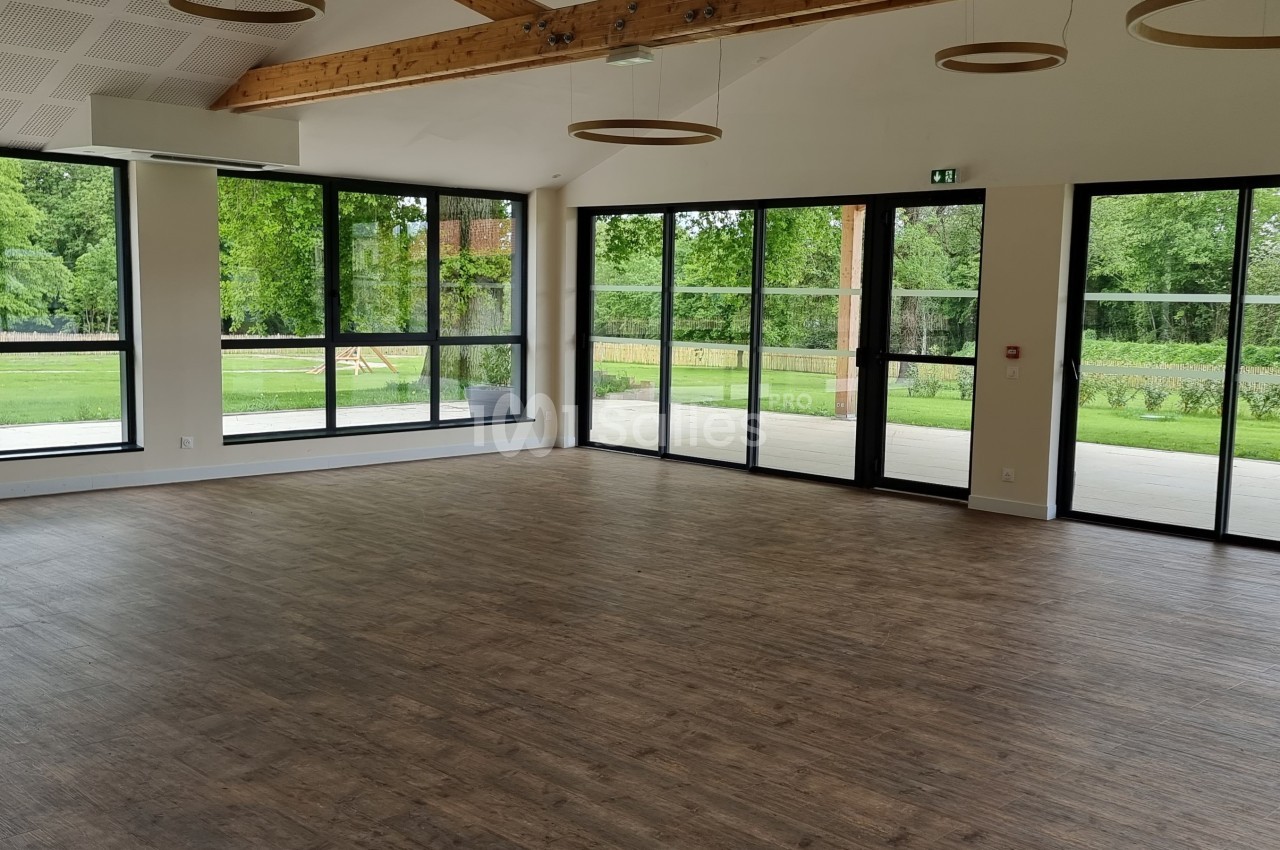 Salle lumineuse avec parquet, grandes baies vitrées donnant sur un jardin verdoyant et poutres apparentes au plafond.