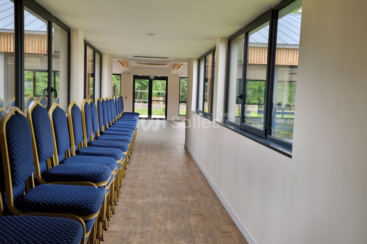 Rangée de chaises bleues alignées le long d'un couloir lumineux avec des fenêtres donnant sur un espace extérieur verdoyant.