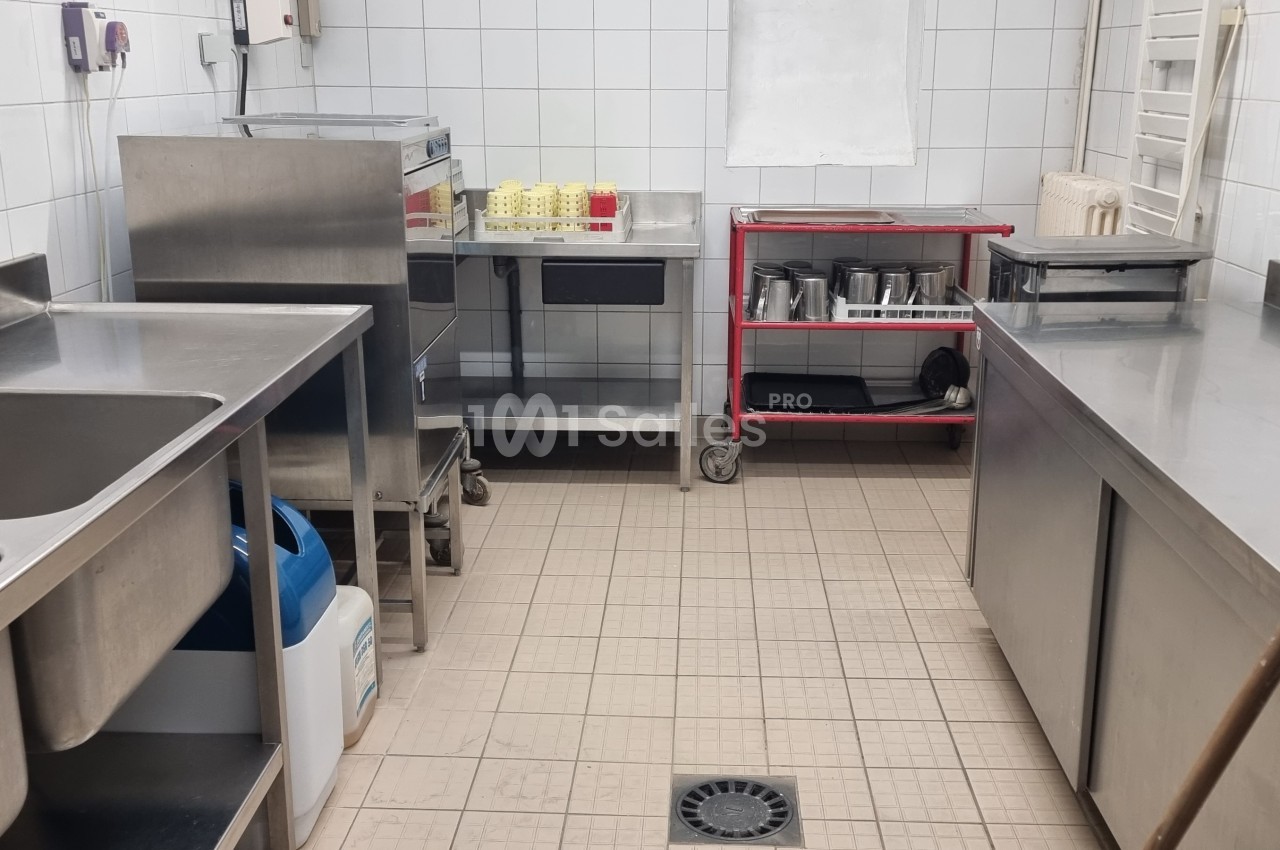Cuisine professionnelle avec équipements en inox, évier double, plan de travail, étagères et sol carrelé beige.